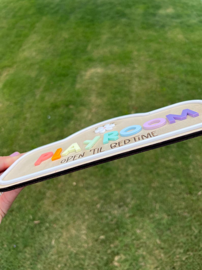 Puede incluir: Un letrero de madera con una superficie clara y un borde oscuro, que presenta la palabra PLAYROOM en letras pastel multicolores en relieve y el texto OPEN TIL BED TIME impreso en negro debajo, con una peque&ntilde;a forma de flor blanca incluida.