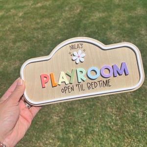 Puede incluir: Un letrero de madera con borde blanco que presenta el texto "MILA'S PLAYROOM" en letras 3D coloridas, un icono de flor blanca y el texto "OPEN 'TIL BEDTIME" impreso en negro. El letrero tiene una forma redondeada similar a una nube.