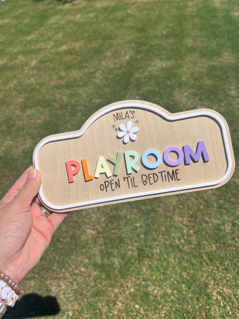 Puede incluir: Un letrero de madera con borde blanco que presenta el texto "MILA'S PLAYROOM" en letras 3D coloridas, un icono de flor blanca y el texto "OPEN 'TIL BEDTIME" impreso en negro. El letrero tiene una forma redondeada similar a una nube.