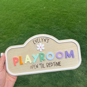 Puede incluir: Un letrero de madera de color claro con un borde blanco que presenta el texto "EVELYN'S" sobre un icono de flor blanca, la palabra "PLAYROOM" en letras 3D multicolores y el texto "OPEN 'TIL BEDTIME" impreso en negro en la parte inferior.