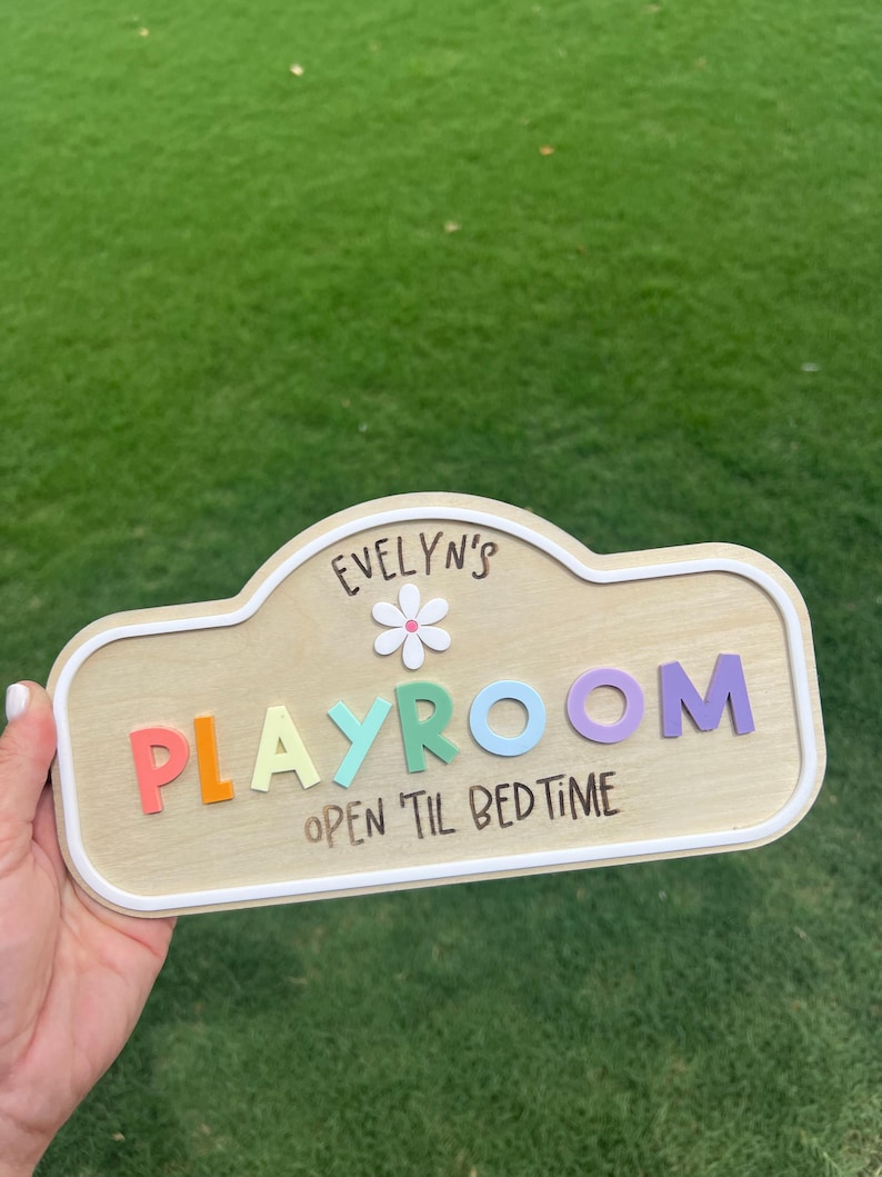 Puede incluir: Un letrero de madera de color claro con un borde blanco que presenta el texto "EVELYN'S" sobre un icono de flor blanca, la palabra "PLAYROOM" en letras 3D multicolores y el texto "OPEN 'TIL BEDTIME" impreso en negro en la parte inferior.
