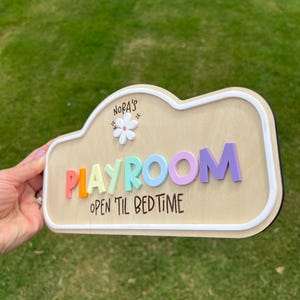 Puede incluir: Un letrero de madera con forma de nube y borde blanco que presenta el texto 'NORA'S' sobre un icono de flor blanca, la palabra 'PLAYROOM' en letras 3D coloridas y el texto 'OPEN 'TIL BEDTIME' impreso en negro sobre la superficie de madera clara.
