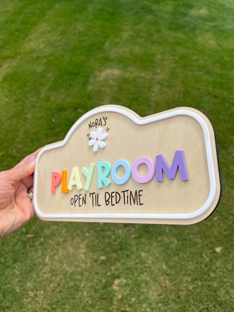 Puede incluir: Un letrero de madera con forma de nube y borde blanco que presenta el texto 'NORA'S' sobre un icono de flor blanca, la palabra 'PLAYROOM' en letras 3D coloridas y el texto 'OPEN 'TIL BEDTIME' impreso en negro sobre la superficie de madera clara.