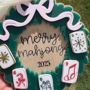 May include: A Christmas wreath-shaped ornament with the words "merry mahjong 2025" in black script. The wreath is green with a pink bow and several small, decorated tiles. The tiles feature festive designs like a snowflake and a candy cane.