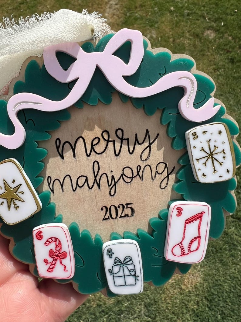 May include: A Christmas wreath-shaped ornament with the words "merry mahjong 2025" in black script. The wreath is green with a pink bow and several small, decorated tiles. The tiles feature festive designs like a snowflake and a candy cane.