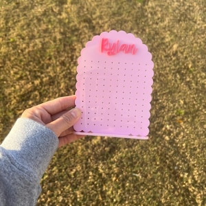 May include: Pink earring holder with a scalloped edge and the name "Rylan" in pink lettering. The holder has multiple holes for earrings.