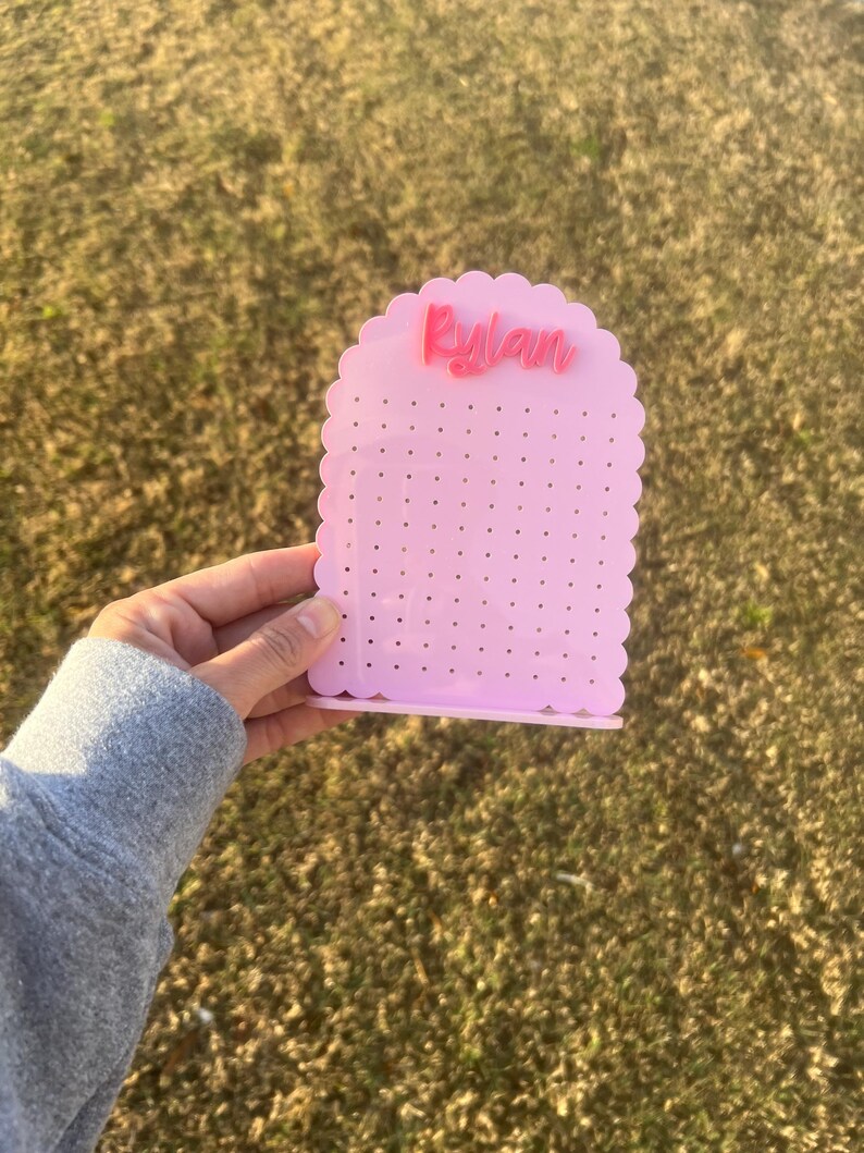 May include: Pink earring holder with a scalloped edge and the name "Rylan" in pink lettering. The holder has multiple holes for earrings.