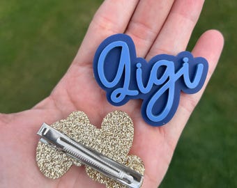 May include: A blue and white name tag that says "Gigi" and a gold glitter hair clip. The name tag is in a cursive font with a blue outline. The hair clip is in the shape of a heart.