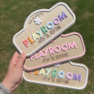 Puede incluir: Tres letreros rectangulares de madera con esquinas redondeadas, cada uno con el texto "PLAYROOM" y "OPEN 'TIL BEDTIME" en letras coloridas en relieve. El letrero superior incluye el texto "LILA & AVERY'S" y un peque&ntilde;o icono de flor blanca.