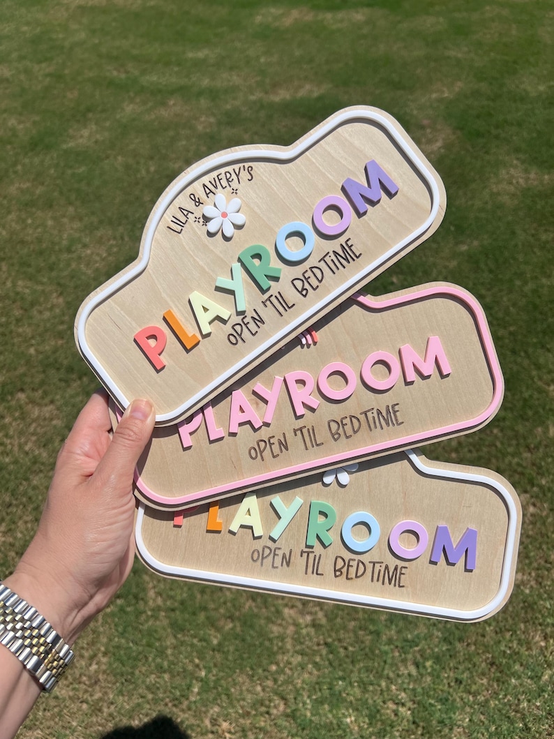 Puede incluir: Tres letreros rectangulares de madera con esquinas redondeadas, cada uno con el texto "PLAYROOM" y "OPEN 'TIL BEDTIME" en letras coloridas en relieve. El letrero superior incluye el texto "LILA & AVERY'S" y un peque&ntilde;o icono de flor blanca.