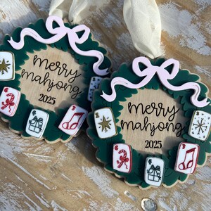 May include: Two Christmas ornaments with a green wreath design, featuring the text "Merry Mahjong 2025". Each ornament has a light pink bow and small, colorful mahjong tile accents. Cream-colored ribbons are attached for hanging.
