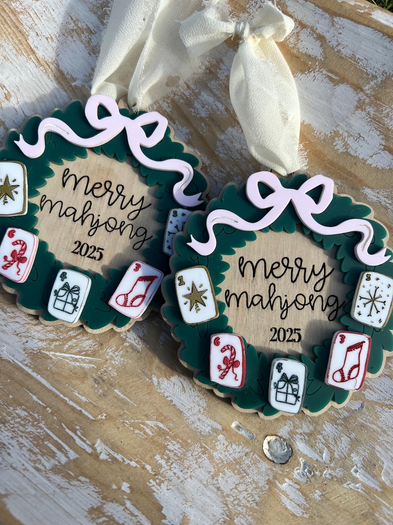 May include: Two Christmas ornaments with a green wreath design, featuring the text "Merry Mahjong 2025". Each ornament has a light pink bow and small, colorful mahjong tile accents. Cream-colored ribbons are attached for hanging.