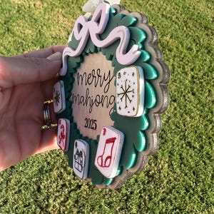May include: A round, layered ornament with a green wreath design and a light pink bow. The ornament has the words "merry mahjong 2025" and several game tiles. The ornament is held in a hand.