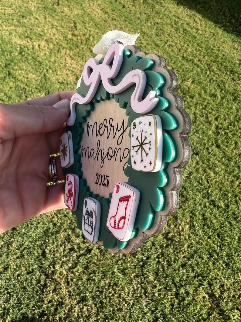 May include: A round, layered ornament with a green wreath design and a light pink bow. The ornament has the words "merry mahjong 2025" and several game tiles. The ornament is held in a hand.