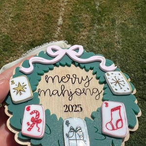 May include: A Christmas ornament with a green wreath design, featuring the words "merry mahjong" and the year 2025. The ornament includes decorative playing cards with festive symbols and a pink bow.