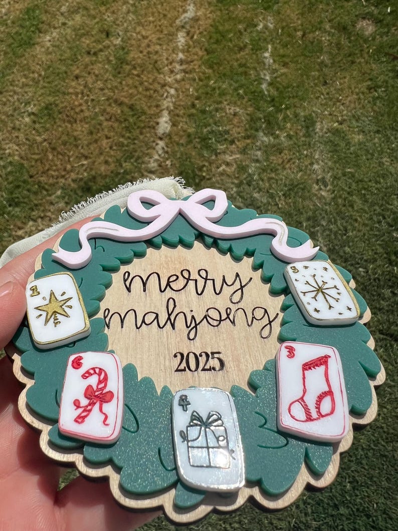 May include: A Christmas ornament with a green wreath design, featuring the words "merry mahjong" and the year 2025. The ornament includes decorative playing cards with festive symbols and a pink bow.