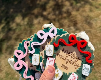 May include: A set of circular wooden ornaments shaped like wreaths with dark green foliage. Each ornament features a colorful bow, small mahjong tile accents with holiday designs, and the engraved text merry mahjong 2026 in the center.