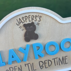 Puede incluir: Un letrero de madera clara con borde blanco que presenta el texto grabado "JASPER'S" sobre un icono de oso de peluche marr&oacute;n. Debajo, letras 3D de color azul brillante forman "PLAYROOM" con el texto grabado "OPEN 'TIL BED TIME".