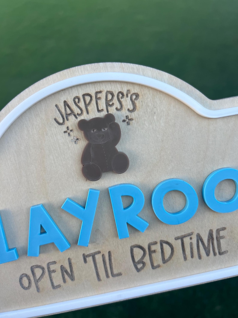 Puede incluir: Un letrero de madera clara con borde blanco que presenta el texto grabado "JASPER'S" sobre un icono de oso de peluche marr&oacute;n. Debajo, letras 3D de color azul brillante forman "PLAYROOM" con el texto grabado "OPEN 'TIL BED TIME".