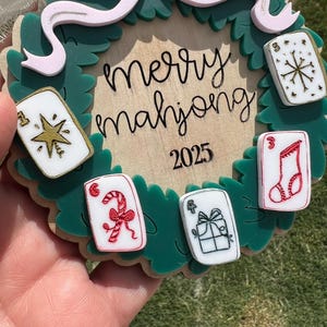 May include: A festive Christmas ornament with a green wreath design, featuring the words "merry mahjong" and the year 2025. The ornament includes decorative mahjong tiles with holiday-themed graphics and a pink bow.