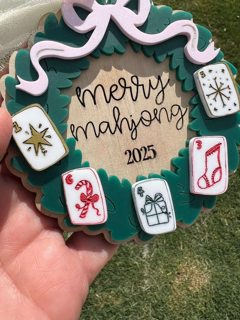 May include: A festive Christmas ornament with a green wreath design, featuring the words "merry mahjong" and the year 2025. The ornament includes decorative mahjong tiles with holiday-themed graphics and a pink bow.