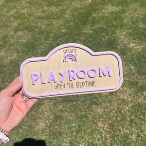 Puede incluir: Un letrero de madera clara con un borde morado y letras moradas en relieve que dicen "BELLA'S" sobre un peque&ntilde;o icono de arco&iacute;ris, seguido de la palabra "PLAYROOM" y el texto "OPEN TIL BEDTIME" en una fuente oscura estilo manuscrito.