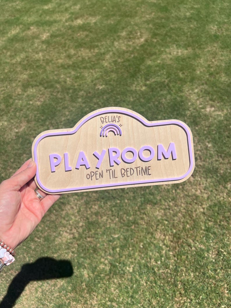 Puede incluir: Un letrero de madera clara con un borde morado y letras moradas en relieve que dicen "BELLA'S" sobre un peque&ntilde;o icono de arco&iacute;ris, seguido de la palabra "PLAYROOM" y el texto "OPEN TIL BEDTIME" en una fuente oscura estilo manuscrito.