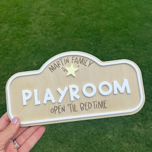 Puede incluir: Un letrero de madera de color claro con un borde blanco en relieve. El texto dice "MARTIN FAMILY" sobre una peque&ntilde;a estrella amarilla, seguido de la palabra "PLAYROOM" en letras blancas grandes y "OPEN 'TIL BEDTIME" en letras oscuras.