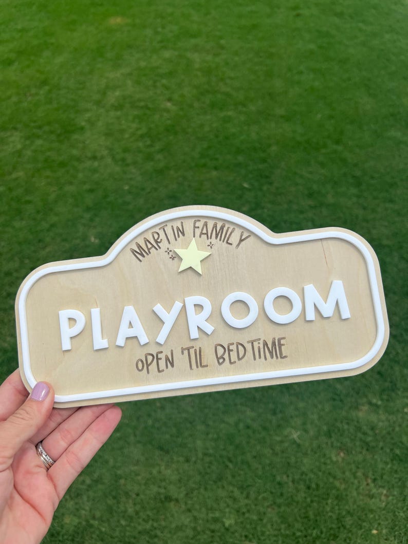 Puede incluir: Un letrero de madera de color claro con un borde blanco en relieve. El texto dice "MARTIN FAMILY" sobre una peque&ntilde;a estrella amarilla, seguido de la palabra "PLAYROOM" en letras blancas grandes y "OPEN 'TIL BEDTIME" en letras oscuras.