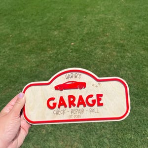 May include: A custom wooden sign with a light wood finish and a red border. It features the text 'HABIB'S GARAGE', a red car silhouette, and the words 'CHECK - REPAIR - ROLL' with 'EST. 2026' printed below in a dark font on the light wood surface.