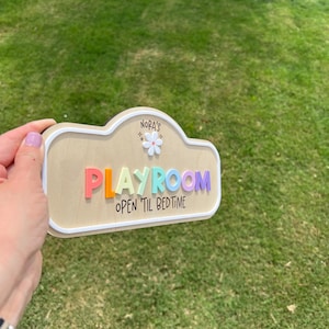 Puede incluir: Un letrero de madera con forma de nube y borde blanco que presenta el texto NORA'S sobre un peque&ntilde;o icono de flor blanca. La palabra PLAYROOM aparece en letras realzadas y multicolores, seguida del texto OPEN 'TIL BEDTIME en negro.