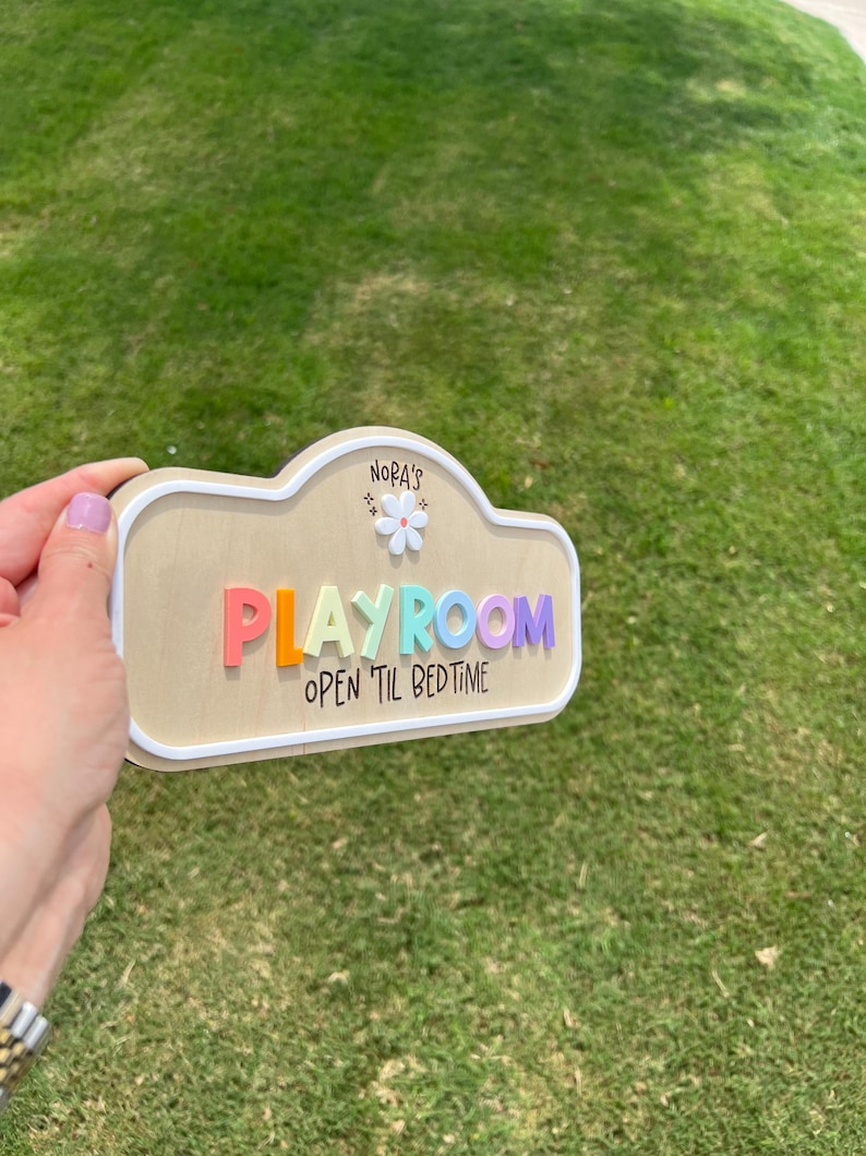 Puede incluir: Un letrero de madera con forma de nube y borde blanco que presenta el texto NORA'S sobre un peque&ntilde;o icono de flor blanca. La palabra PLAYROOM aparece en letras realzadas y multicolores, seguida del texto OPEN 'TIL BEDTIME en negro.