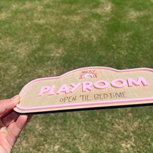 Puede incluir: Un letrero de madera con forma de nube, acabado en madera clara y borde rosa en relieve. El letrero muestra el texto "MILA'S" sobre un peque&ntilde;o gr&aacute;fico de arco&iacute;ris, la palabra "PLAYROOM" en letras rosas grandes y "OPEN 'TIL BED TIME" en oscuro.