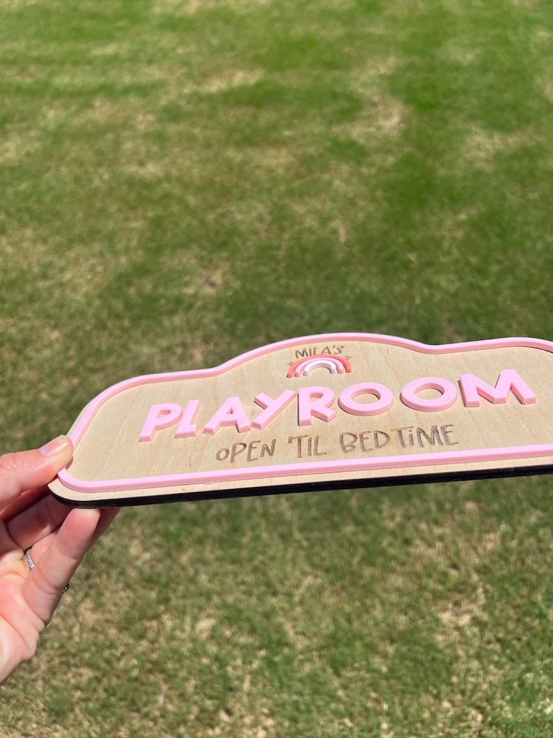 Puede incluir: Un letrero de madera con forma de nube, acabado en madera clara y borde rosa en relieve. El letrero muestra el texto "MILA'S" sobre un peque&ntilde;o gr&aacute;fico de arco&iacute;ris, la palabra "PLAYROOM" en letras rosas grandes y "OPEN 'TIL BED TIME" en oscuro.