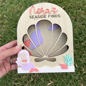 May include: Wooden mermaid-themed display with the text "Nora's Seaside Finds". Features a mermaid, seashells, and coral accents. The display has a seashell-shaped cutout and a white base.