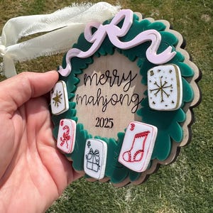 May include: A green and brown Christmas ornament with a wreath design. The ornament features the text "merry mahjong 2025" and several small, colorful mahjong tiles. A pink bow and white ribbon are attached for hanging.