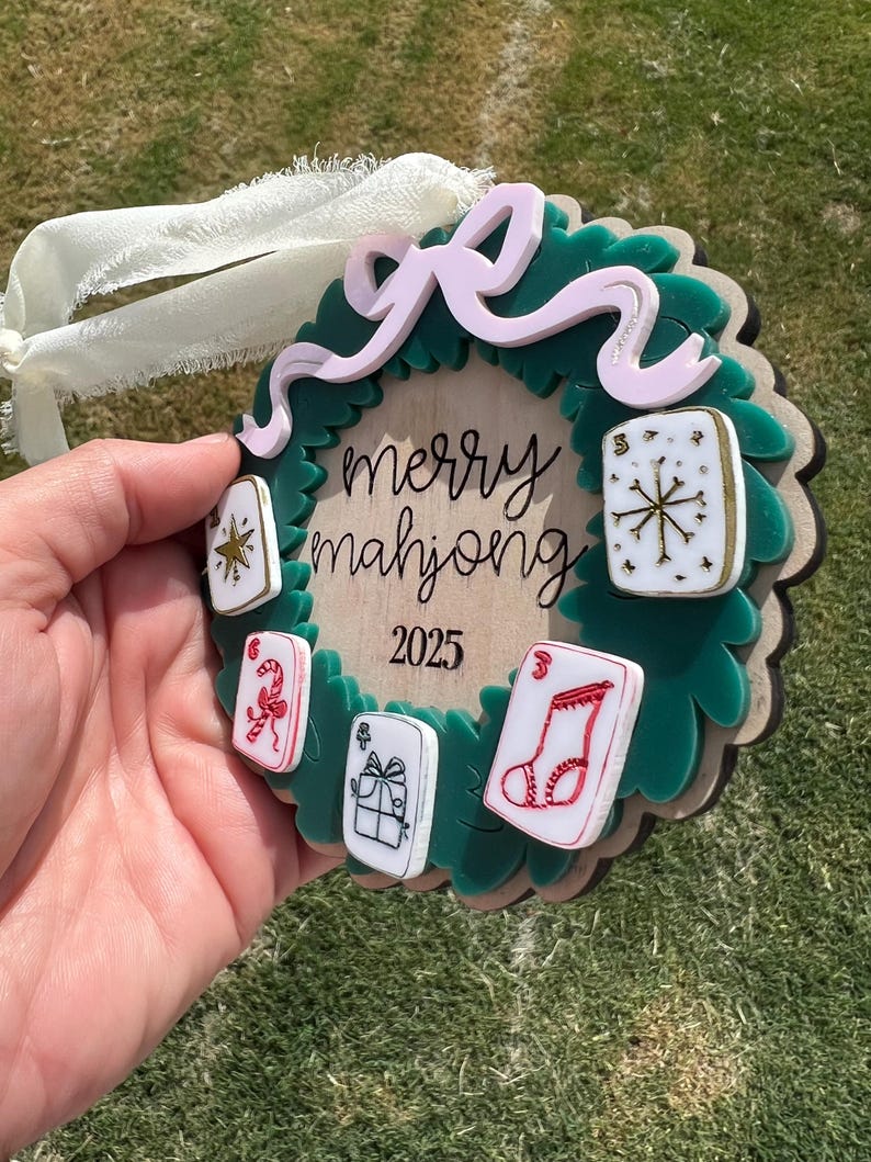 May include: A green and brown Christmas ornament with a wreath design. The ornament features the text "merry mahjong 2025" and several small, colorful mahjong tiles. A pink bow and white ribbon are attached for hanging.