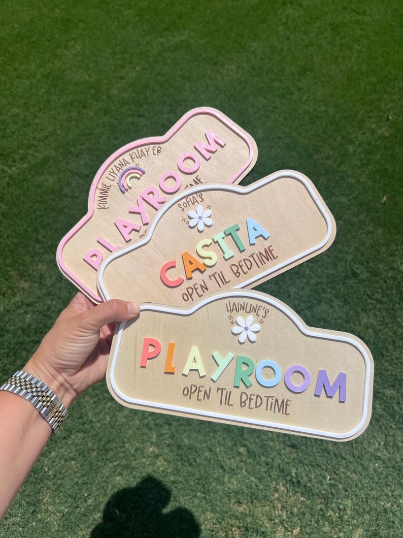 Puede incluir: Tres letreros de madera personalizados con bordes redondeados y letras coloridas en relieve. Los letreros incluyen los textos KIMMIE LIYANA KHAYER PLAYROOM, SOFIA'S CASITA OPEN 'TIL BEDTIME y HAINLINE'S PLAYROOM OPEN 'TIL BEDTIME, decorados con iconos de arco&iacute;ris y flores.