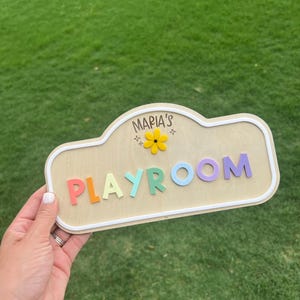 Puede incluir: Un letrero de madera de color claro con un borde blanco, que presenta el texto "MARIA'S" sobre un icono de flor amarilla y la palabra "PLAYROOM" en letras de bloque elevadas y multicolores en rojo, naranja, verde claro, azul y morado.