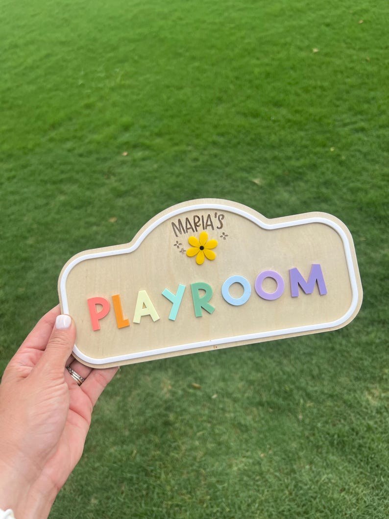 Puede incluir: Un letrero de madera de color claro con un borde blanco, que presenta el texto "MARIA'S" sobre un icono de flor amarilla y la palabra "PLAYROOM" en letras de bloque elevadas y multicolores en rojo, naranja, verde claro, azul y morado.