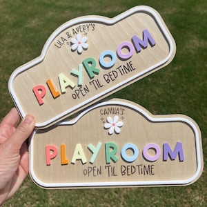 Puede incluir: Dos letreros de madera personalizados con fondo de veta de madera clara y bordes blancos en relieve. Cada letrero presenta una flor blanca, la palabra 'PLAYROOM' en letras 3D multicolores y el texto negro 'OPEN 'TIL BEDTIME' con nombres.
