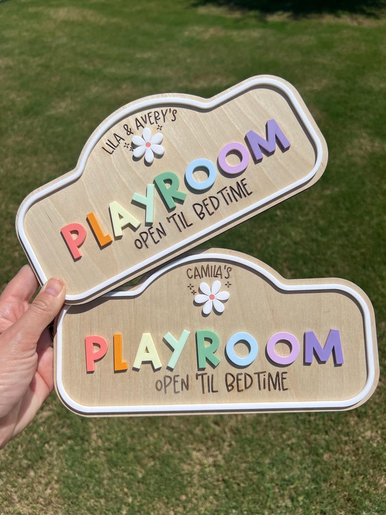 Puede incluir: Dos letreros de madera personalizados con fondo de veta de madera clara y bordes blancos en relieve. Cada letrero presenta una flor blanca, la palabra 'PLAYROOM' en letras 3D multicolores y el texto negro 'OPEN 'TIL BEDTIME' con nombres.