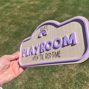 Puede incluir: Un letrero de madera de color claro con un borde morado en relieve y letras moradas en 3D que dicen "BELLA'S" sobre un peque&ntilde;o icono de arco&iacute;ris, seguido de la palabra "PLAYROOM" y el texto "OPEN TIL BED TIME" en fuente negra.