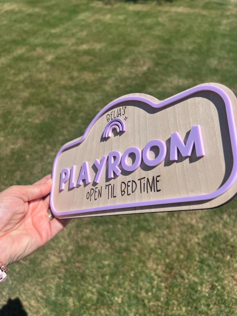 Puede incluir: Un letrero de madera de color claro con un borde morado en relieve y letras moradas en 3D que dicen "BELLA'S" sobre un peque&ntilde;o icono de arco&iacute;ris, seguido de la palabra "PLAYROOM" y el texto "OPEN TIL BED TIME" en fuente negra.