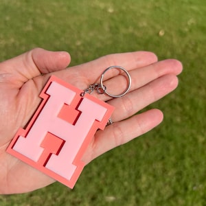 May include: A pink and coral letter "H" keychain. The keychain features a layered design with a light pink center and a coral outline. The metal keyring is attached to the top of the letter.