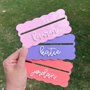 May include: Three colorful place cards in pink, purple, and coral, each with a scalloped edge and a slot to hold a name. The names "Lauren", "Katie", and "Jordan" are written in white script.