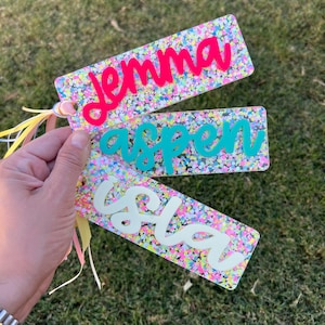 May include: Three rectangular name tags with colorful confetti backgrounds. The names "Jemma" in hot pink, "Aspen" in teal, and "Isla" in white are displayed in a cursive font. Each tag has a ribbon attached.