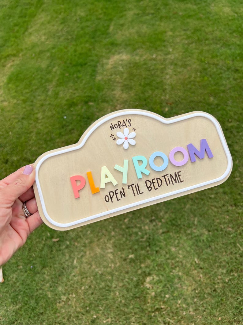 Puede incluir: Un letrero de madera con borde blanco que presenta el texto "NORA'S" sobre un icono de flor blanca, la palabra "PLAYROOM" en letras coloridas en relieve y el texto "OPEN 'TIL BEDTIME" impreso en negro en la parte inferior del letrero.