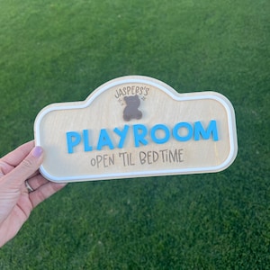 Puede incluir: Un letrero de madera de color claro con un borde blanco que presenta el texto 'JASPERS'S' sobre un peque&ntilde;o icono de oso marr&oacute;n, la palabra 'PLAYROOM' en letras azules en relieve y el texto 'OPEN 'TIL BEDTIME' en fuente oscura.