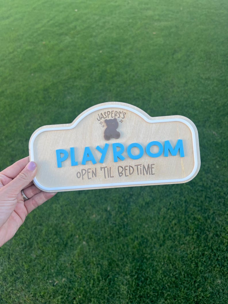 Puede incluir: Un letrero de madera de color claro con un borde blanco que presenta el texto 'JASPERS'S' sobre un peque&ntilde;o icono de oso marr&oacute;n, la palabra 'PLAYROOM' en letras azules en relieve y el texto 'OPEN 'TIL BEDTIME' en fuente oscura.